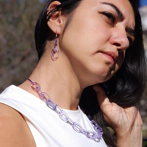 May include: A clear, lavender-colored chain necklace and matching earrings. The necklace is composed of interlocking oval links, and the earrings have a similar design. The jewelry is worn with a white top.
