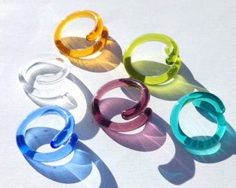 May include: A collection of colorful glass rings in shades of yellow, green, clear, purple, blue, and teal. Each ring has a spiral design, casting shadows on a white surface. The rings are a unique accessory.