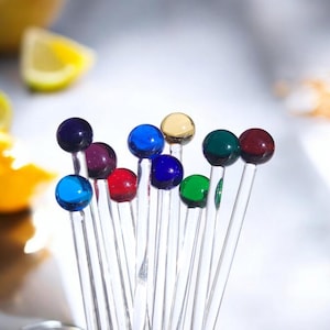 Peut inclure: Un ensemble de dix bâtonnets de mélange en verre avec des perles de verre colorées sur le dessus. Les perles sont de différentes couleurs, notamment le bleu, le vert, le rouge, le violet et le jaune.