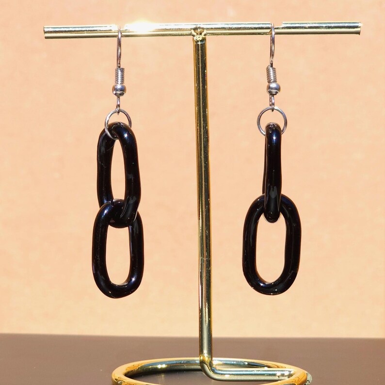 May include: A pair of black chain link earrings. The earrings are displayed on a gold-colored jewelry stand. The chain links are glossy and hang from silver-colored ear wires.
