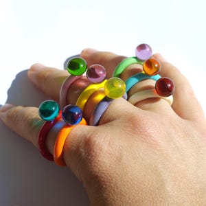 Peut inclure: Une main ornée de plusieurs bagues colorées. Chaque bague présente une bande de couleur différente, notamment rouge, orange, jaune, vert, bleu et violet. Chaque bague est surmontée d'une petite sphère de couleur assortie. Les bagues sont en matériau brillant.