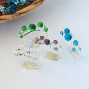 Peut inclure: Quatre bagues en résine transparente ornées de petites sphères en relief de différentes couleurs. Une bague présente des sphères vertes, une des sphères bleues, une des sphères violettes marbrées et une des sphères transparentes.