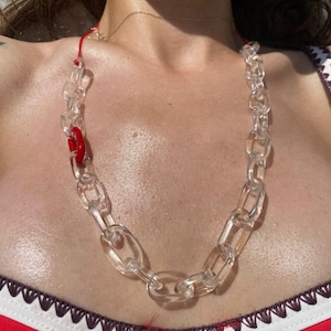 Könnte beinhalten: Eine transparente Acryl-Kettenhalskette mit großen ovalen Gliedern. Die Halskette hat ein rotes Akzentstück und eine rote Kordel. Die Halskette wird über einem roten Oberteil mit weißem Besatz getragen.