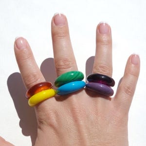 May include: A hand displays six colorful, oval-shaped rings. The rings are stacked in pairs, with colors including amber and yellow, green and blue, and purple and black. The rings are made of a glossy material.