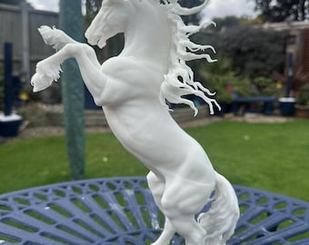 May include: A white sculpture of a rearing horse. The horse is sculpted with detailed features, including a flowing mane and tail. The sculpture is set on a blue metal table, with a blurred outdoor background.