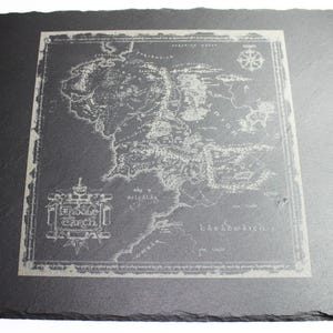 May include: A square, dark gray slate coaster featuring a detailed map of Middle-earth, with the words "Middle Earth" and other text. The map is etched in a lighter gray, creating a contrast against the darker slate.