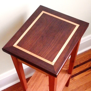 May include: A small wooden end table with a dark wood top and a lighter wood shelf. The top has a decorative border of light wood.