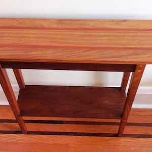 May include: A wooden end table with a curved top and a lower shelf. The top is made of light wood with darker wood inlay. The legs are made of dark wood.
