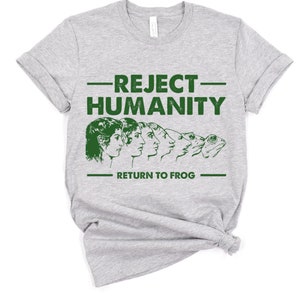 May include: A light gray t-shirt with the text "REJECT HUMANITY" above a line of evolving faces transforming into a frog, with the text "RETURN TO FROG" below. The graphic is printed in green.
