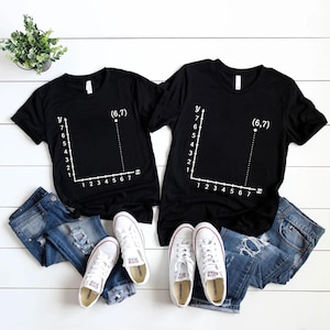 May include: Two black t-shirts with a white graph design and the coordinates (6,7). The shirts are paired with blue jeans and white sneakers. A small green plant in a white pot is visible in the background.