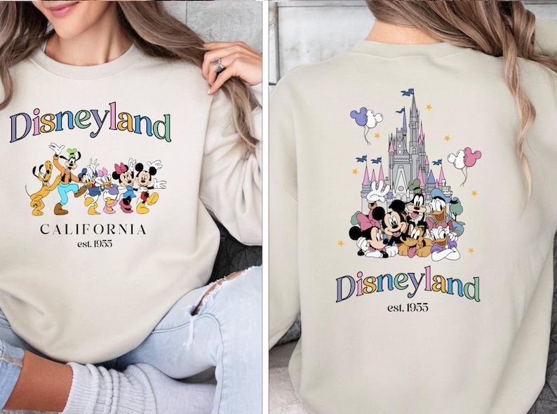 Puede incluir: Una sudadera de color crema con un gr&aacute;fico de "Disneyland" colorido en la parte delantera. El gr&aacute;fico presenta a Mickey Mouse, Minnie Mouse, Goofy, Donald Duck y Pluto. El texto "California est. 1955" est&aacute; debajo del gr&aacute;fico. La parte posterior de la sudadera presenta un gr&aacute;fico colorido del castillo de la Bella Durmiente con Mickey Mouse, Minnie Mouse, Goofy, Donald Duck y Pluto en frente. El texto "Disneyland est. 1955" est&aacute; debajo del gr&aacute;fico.