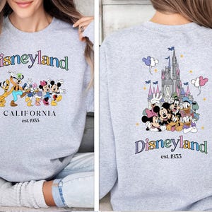 Puede incluir: Sudadera gris con un logotipo de Disneyland colorido y personajes de Mickey Mouse en la parte delantera. La parte posterior de la sudadera presenta una ilustraci&oacute;n colorida del castillo de la Bella Durmiente con Mickey Mouse y sus amigos delante. El texto "Disneyland est. 1955" est&aacute; impreso en la parte posterior.