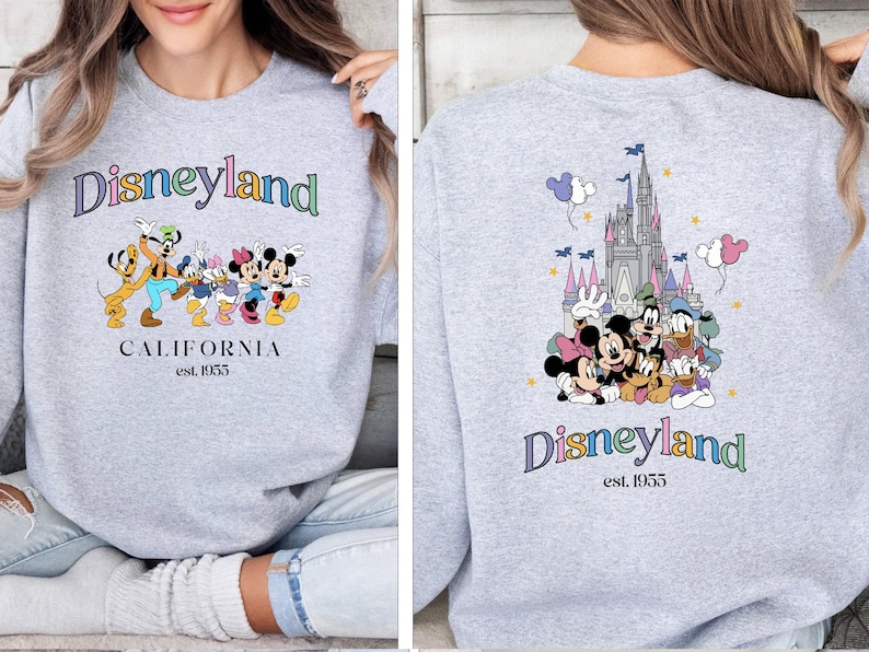 Puede incluir: Sudadera gris con un logotipo de Disneyland colorido y personajes de Mickey Mouse en la parte delantera. La parte posterior de la sudadera presenta una ilustraci&oacute;n colorida del castillo de la Bella Durmiente con Mickey Mouse y sus amigos delante. El texto "Disneyland est. 1955" est&aacute; impreso en la parte posterior.