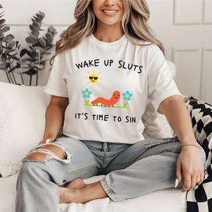 May include: White t-shirt with the text "WAKE UP SLUTS IT'S TIME TO SIN". The graphic includes a smiling sun wearing sunglasses, a red worm, and blue flowers. The shirt is worn by a person.