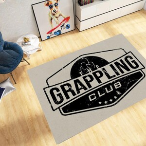 May include: A rectangular rug with a distressed black and white logo that reads "The Grappling Club." The rug is light grey and has a worn appearance. The logo features a silhouette of two figures grappling.