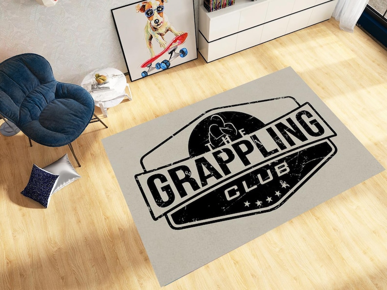 May include: A rectangular rug with a distressed black and white logo that reads "The Grappling Club." The rug is light grey and has a worn appearance. The logo features a silhouette of two figures grappling.