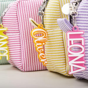 Może przedstawiać: Zbliżenie na trzy małe, pasiasto-tkaninowe torby w kolorze różowym, żółtym i fioletowym. Każda torba ma srebrny zamek błyskawiczny i kolorową etykietę z imieniem: "Oliver" w kolorze pomarańczowym i "Leona" w kolorze różowym. Torby to prawdopodobnie małe torebki lub kosmetyczki.