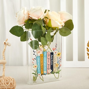 Puede incluir: Un jarrón rectangular transparente con un diseño floral y títulos de libros clásicos. El jarrón contiene rosas de color rosa claro y hojas verdes. Los títulos incluyen "Pride and Prejudice" y "Little Women."