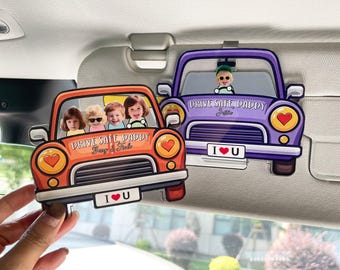 May include: Two car-shaped sun visor ornaments. One is orange with four children's faces, the other is purple with a child's face. Both have the text "DRIVE SAFE, DADDY" and "I ❤️ U".