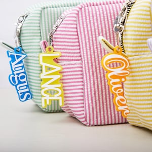 May include: Close-up of three small, striped cosmetic bags in pastel colors: green, pink, and yellow. Each bag has a matching name tag that reads "August", "Lance", and "Oliver". The bags have silver zippers.
