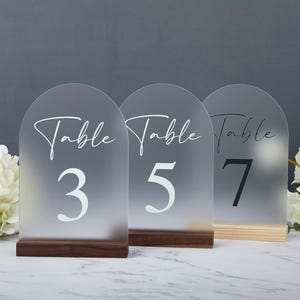 Puede incluir: Tres letreros de número de mesa de acrílico esmerilado con base de madera. Los letreros están numerados 3, 5 y 7. Cada letrero tiene la palabra "Mesa" escrita en cursiva sobre el número.