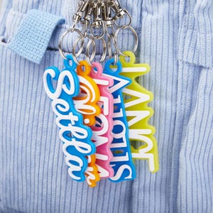 Puede incluir: Una colección de llaveros coloridos con nombres en fuente cursiva. Los llaveros son azules, naranjas, rosas, azules y amarillos, cada uno con un contorno blanco. Están unidos a anillos de metal plateado.