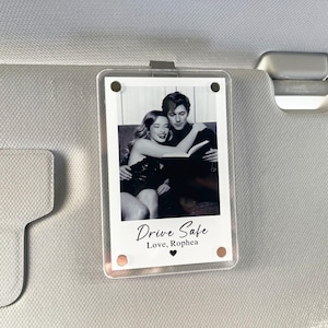 Peut inclure: Support photo en acrylique transparent avec une photo en noir et blanc d'un couple lisant un livre. Le texte "Drive Safe, Love, Rophea" est imprimé en dessous. Le support est fixé à un pare-soleil de voiture.