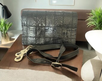 May include: Black rectangular handbag with a crocodile skin pattern. The bag has a flap closure and a detachable black shoulder strap with gold-coloured hardware. The bag is on a wooden surface.