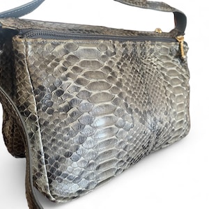 Puede incluir: Bolso bandolera de piel de serpiente gris y blanco con una correa larga y ajustable. El bolso tiene un cierre de cremallera y un tirador dorado. El estampado de piel de serpiente es detallado y texturizado, dando al bolso un aspecto lujoso.