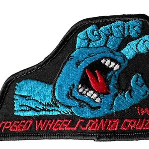 May include: A black and blue embroidered patch with a screaming hand and the text "Speed Wheel Santa Cruz" in red.