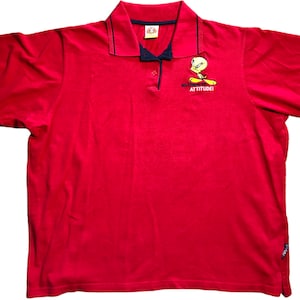 May include: Red polo shirt featuring a Tweety Bird graphic with the text "WHAT'S UP ATTITUDE!". The shirt has a navy blue collar and trim. The short-sleeved shirt is made of a soft, comfortable material.