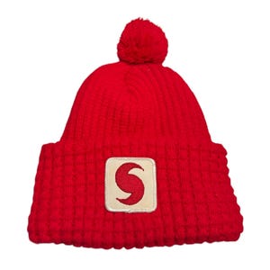 May include: Red knit beanie with a white pom pom on top and a red and white patch with a stylized 'S' on the front.