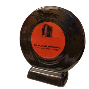 May include: Black ceramic record-shaped coaster with a red label featuring a building silhouette and the text "The Westin Bonaventure LA's Landmark Hotel".