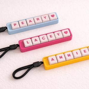 May include: Three personalised keychains in blue, pink, and yellow. Each keychain features a name spelled out with white lettered keys and a pink heart symbol. Black wrist straps are attached to each keychain.