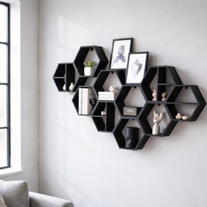 Peut inclure: Étagère murale noire en forme de nid d'abeille. Les étagères sont remplies d'objets décoratifs, notamment de petites plantes, des œuvres d'art encadrées, des livres et des figurines. L'unité est fixée à un mur blanc.