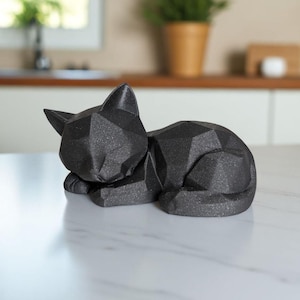 Modern black 3D printed low poly cat sculpture in a sleeping pose on a minimalist desk with art supplies and a bust in the background