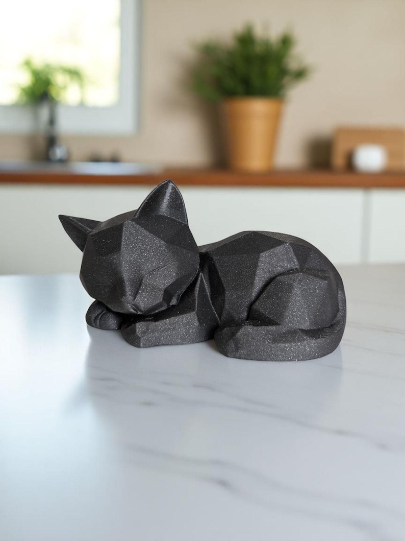 Modern black 3D printed low poly cat sculpture in a sleeping pose on a minimalist desk with art supplies and a bust in the background