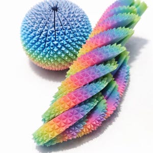 Puede incluir: Un conjunto de juguetes sensoriales fidget de colores. El conjunto incluye una bola puntiaguda y un juguete segmentado flexible, ambos en un degradado arcoíris. Los juguetes tienen una superficie texturizada y están diseñados para la estimulación táctil. El texto "SHOP NOW!" está incluido.