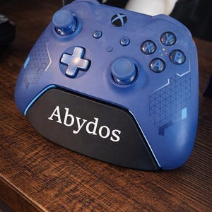 以下が含まれることがあります： 黒いベースに白い文字で「Abydos」と書かれた青いXboxコントローラー。 コントローラーには青いボタンと方向パッドがあります。 コントローラーは木製の表面にあります。