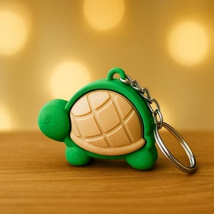 May include: A green turtle keychain with a light brown shell and silver chain. The turtle has a detailed shell pattern and a loop for attaching keys. The keychain is made of a smooth material and sits on a wooden surface.