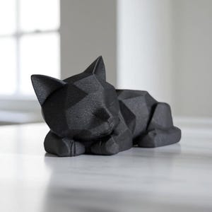 Modern black 3D printed low poly cat sculpture in a sleeping pose on a minimalist desk with art supplies and a bust in the background