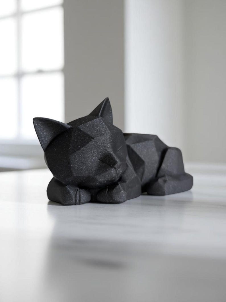 Modern black 3D printed low poly cat sculpture in a sleeping pose on a minimalist desk with art supplies and a bust in the background
