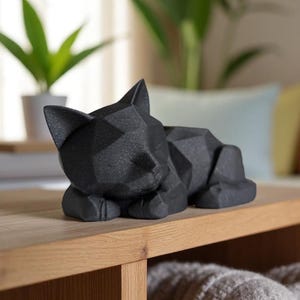 Modern black 3D printed low poly cat sculpture in a sleeping pose on a minimalist desk with art supplies and a bust in the background