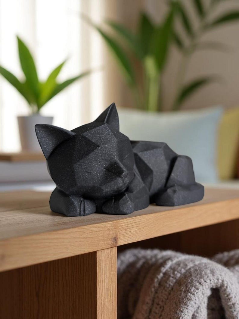 Modern black 3D printed low poly cat sculpture in a sleeping pose on a minimalist desk with art supplies and a bust in the background