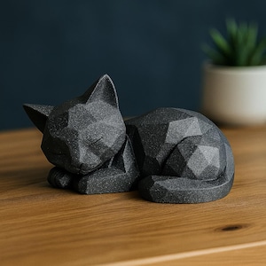 Modern black 3D printed low poly cat sculpture in a sleeping pose on a minimalist desk with art supplies and a bust in the background