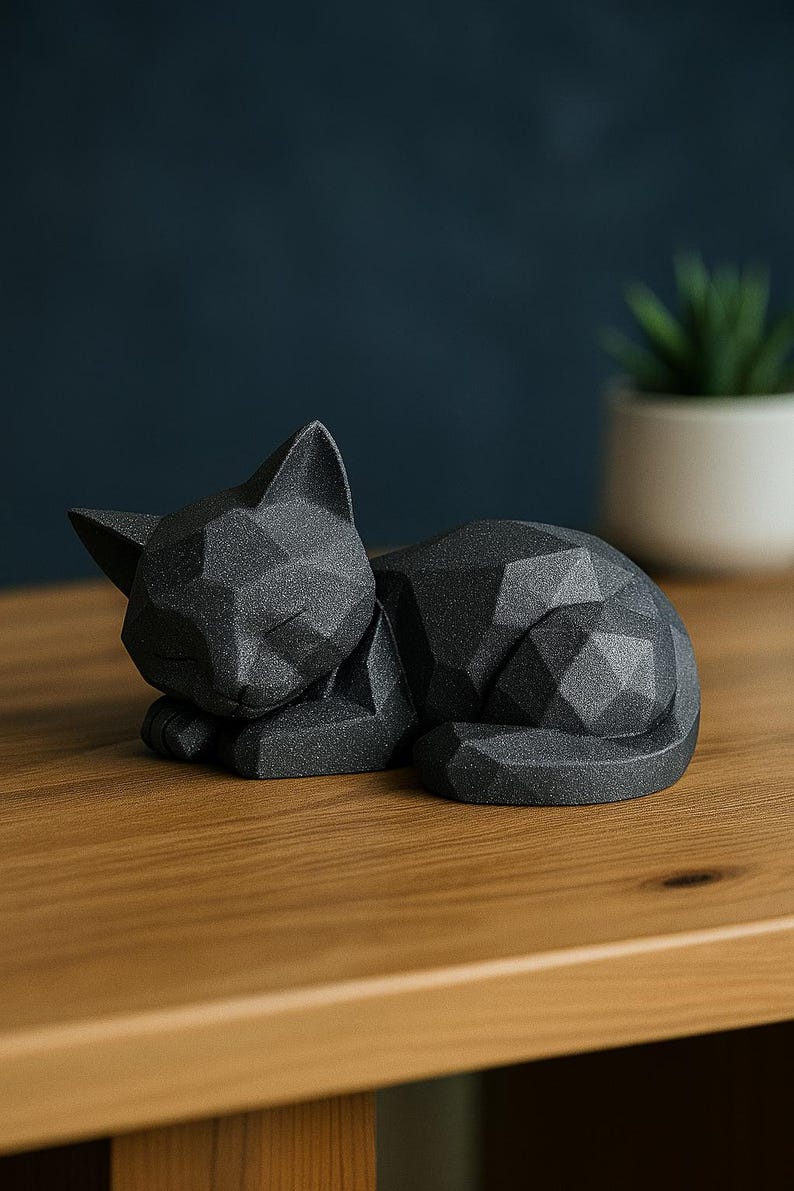 Modern black 3D printed low poly cat sculpture in a sleeping pose on a minimalist desk with art supplies and a bust in the background