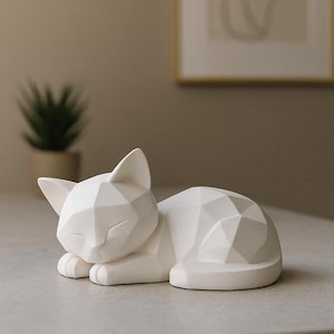 Modern white 3D printed low poly cat sculpture in a sleeping pose on a minimalist desk with art supplies and a bust in the background