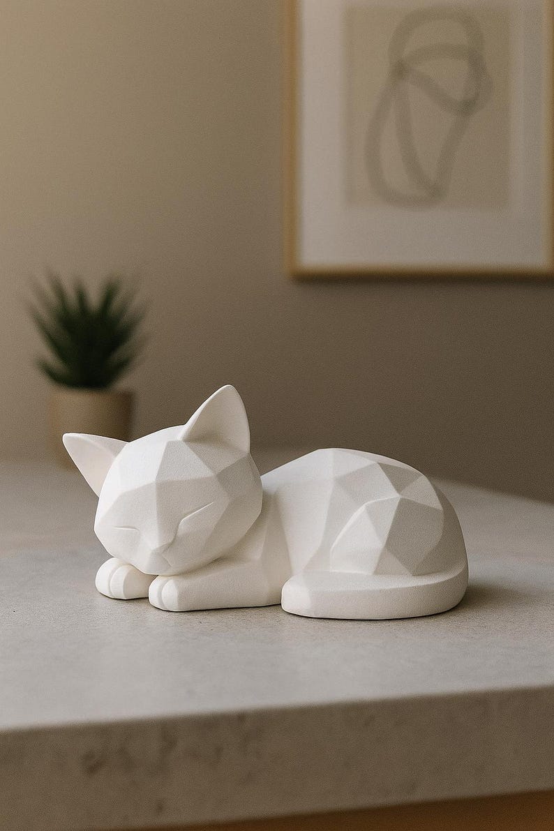 Modern white 3D printed low poly cat sculpture in a sleeping pose on a minimalist desk with art supplies and a bust in the background