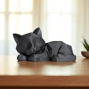 Modern black 3D printed low poly cat sculpture in a sleeping pose on a minimalist desk with art supplies and a bust in the background