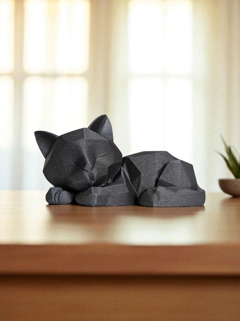 Modern black 3D printed low poly cat sculpture in a sleeping pose on a minimalist desk with art supplies and a bust in the background